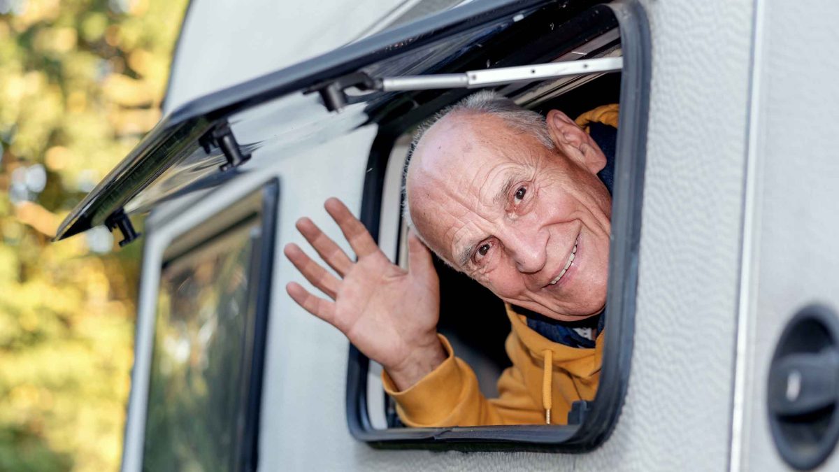 Ein älterer Mann winkt aus dem geöffneten Fenster eines Wohnmobils.